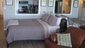 A bedroom with a large bed, a brown leather sofa, a small table, and a kitchen area visible through a window. - Beachfront Gulf View Retreat on Sand Key Beach (Clearwater)
