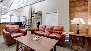 A spacious living room with a brown leather sofa, a wooden coffee table, and a lamp. A staircase leads to another level, and a door is visible in the background. - Park City Resort Ctr 3BR 2Ba Walk2Ski; Free bus Main St; Hot Tub; Winter Pool (Park City)