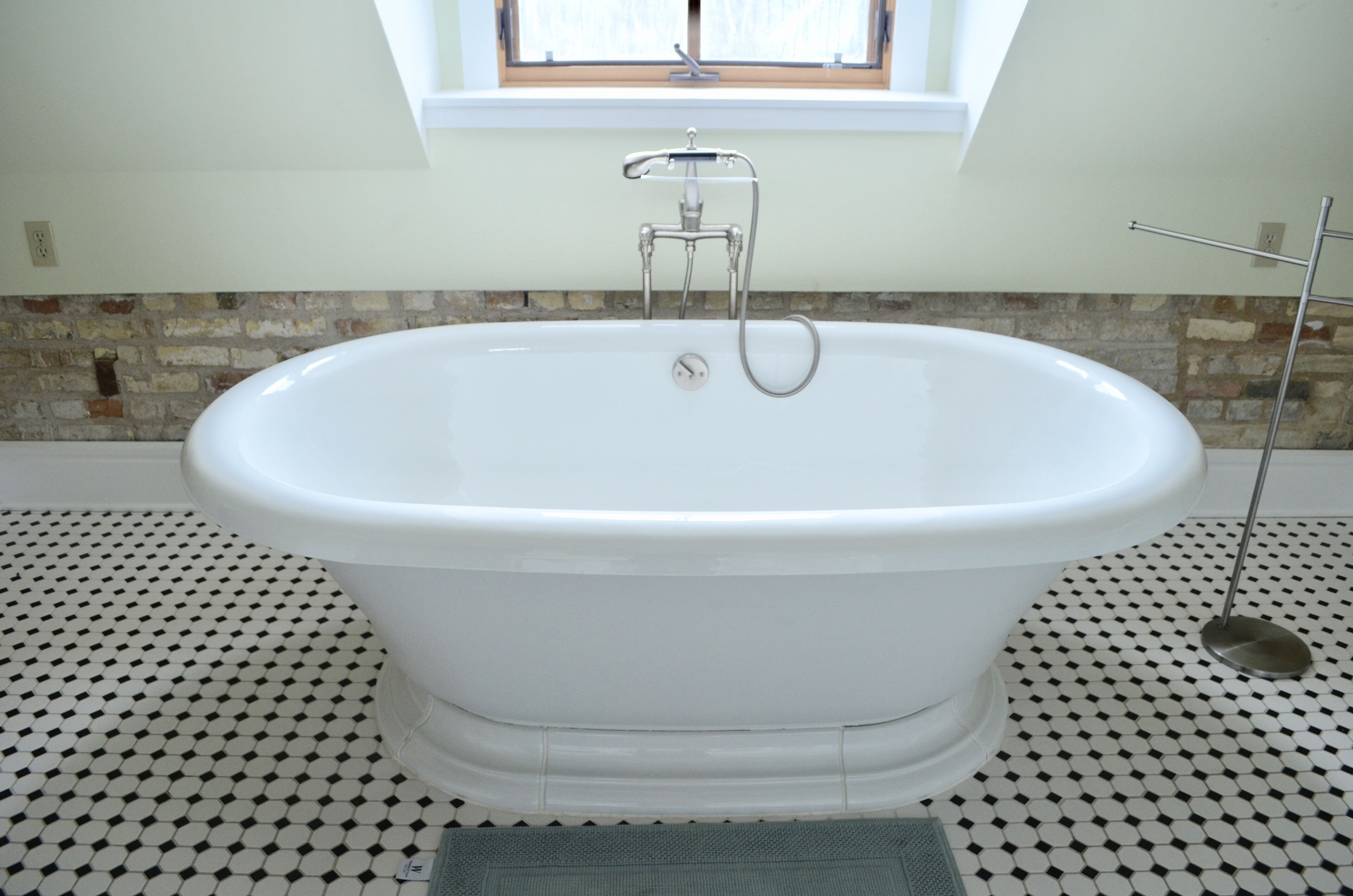 Eine freistehende weiße Badewanne mit Duschbrause, vor einem gefliesten Boden mit geometrischem Muster.
