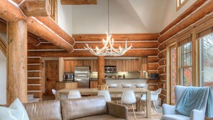 A log cabin interior with a leather sofa, a dining table with chairs, a kitchen area, and a large window.