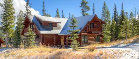 A log cabin with a balcony, surrounded by tall pine trees and a snowy landscape.