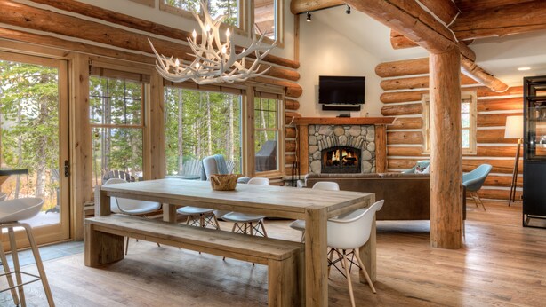 A spacious log cabin interior with a large dining table, multiple chairs, a stone fireplace, and large windows offering a view of the surrounding forest.