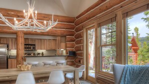 A rustic cabin interior with a wooden dining area, white chairs, a chandelier, and large windows offering a view of the surrounding forest.