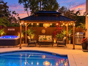 Un patio trasero con un jacuzzi, mobiliario de jardín, una pérgola con luces colgantes, una parrilla y una piscina.