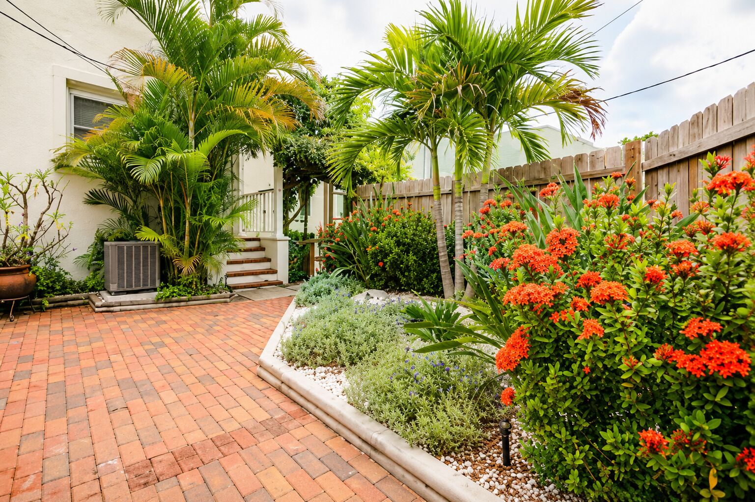 Un patio empedrado con cerca de madera, un edificio blanco y diversas plantas y flores.