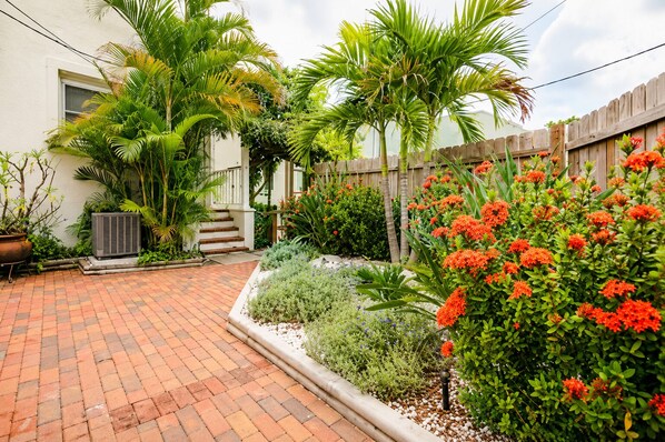 A paved patio with a wooden fence, a white building, and various plants and flowers.
