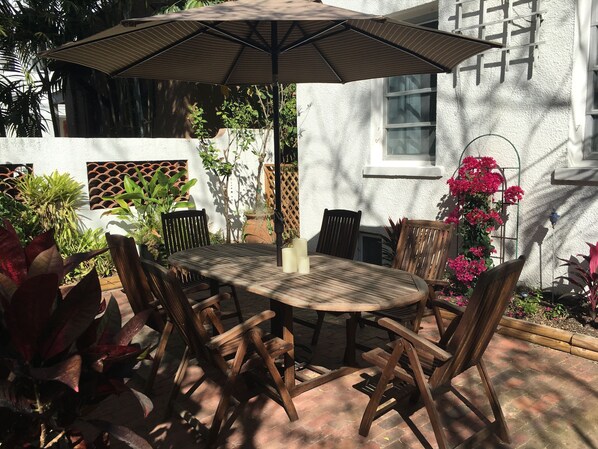 Eine Terrasse im Freien mit einem runden Holztisch und Stühlen, einem Sonnenschirm und verschiedenen Topfpflanzen.