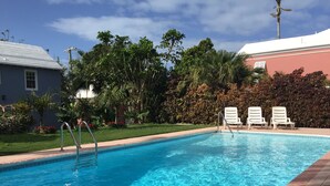Ein Swimmingpool mit klarem, blauem Wasser, umgeben von einem Garten mit verschiedenen Pflanzen und Bäumen. Weiße Liegestühle befinden sich entlang des Poolbereichs.