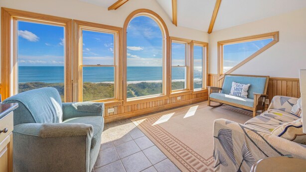 Una habitación amplia con ventanas grandes que ofrecen vistas al océano, una zona de estar acogedora con sofá y sillones, y paneles de madera en las paredes.