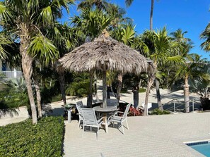 A thatched-roof gazebo with outdoor seating, surrounded by palm trees and a pool area.