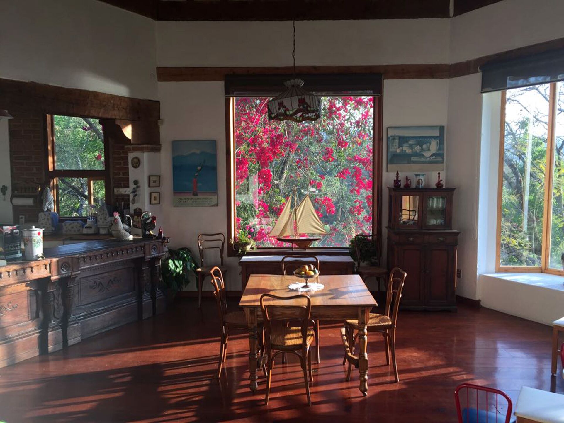 A cozy dining area with a wooden table and chairs, a window with a view of pink flowers, and a rustic kitchen with a brick wall and wooden beams.