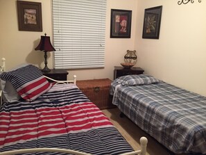 Une chambre avec deux lits, une fenêtre avec des stores, une lampe et des tableaux encadrés accrochés au mur.