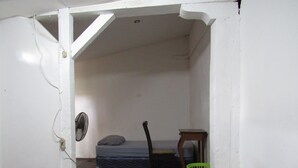 Una stanza piccola e arredata in modo essenziale, con un letto singolo, una sedia, un tavolino e una ventola.