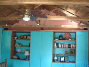 Une pièce dotée d’un plafond en bois et de poutres apparentes, d’un ventilateur de plafond et de deux bibliothèques peintes en turquoise, remplies de livres, d’objets décoratifs et de photos encadrées.