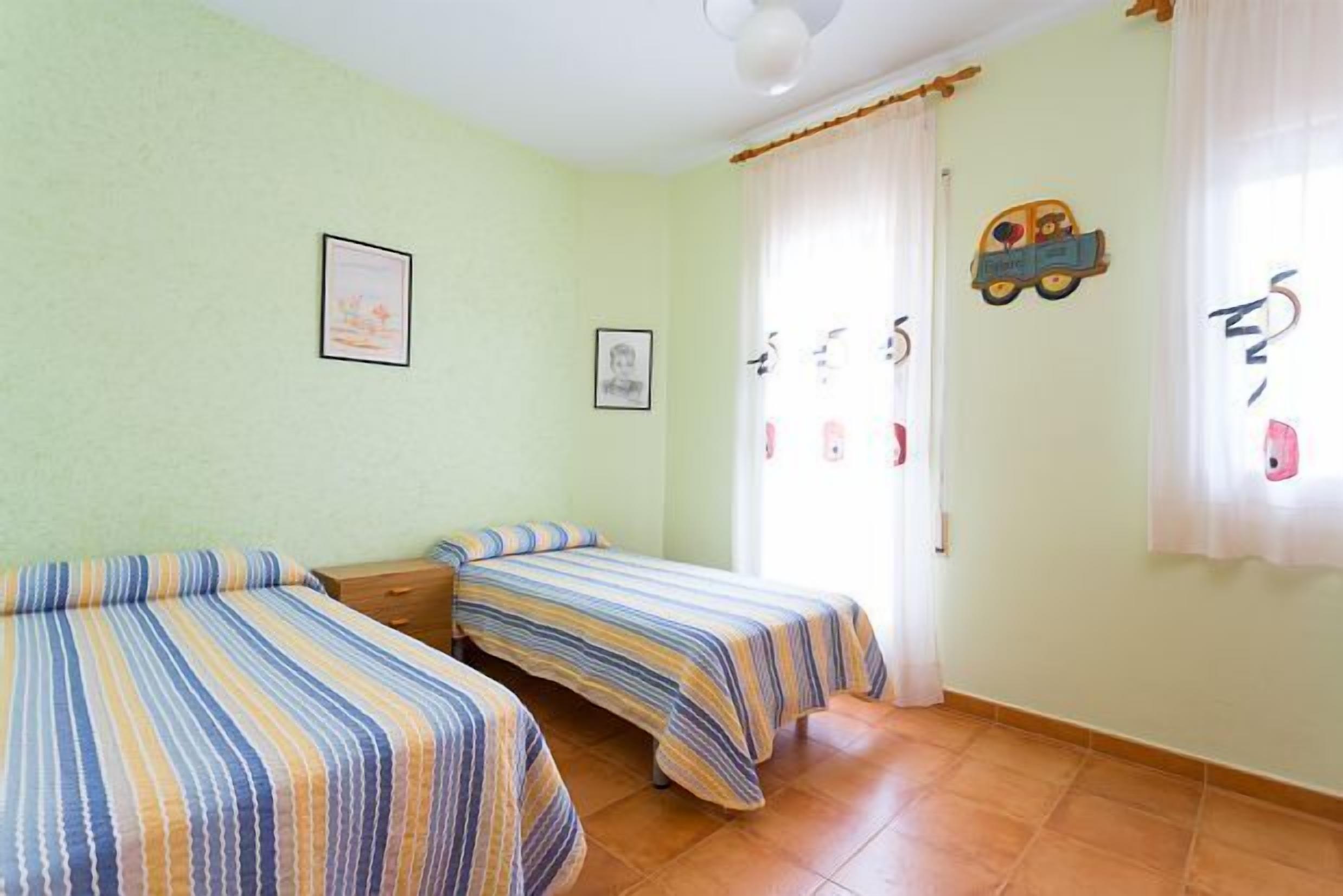 Una habitación con dos camas individuales, una mesita de noche de madera, una ventana con cortinas y adornos en la pared, incluyendo cuadros y un adorno con forma de coche.