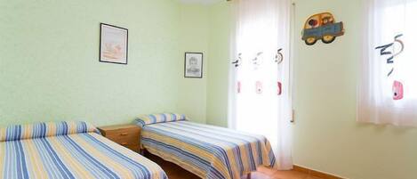 Una habitación con dos camas individuales, una mesita de noche de madera, una ventana con cortinas y adornos en la pared, incluyendo cuadros y un adorno con forma de coche.