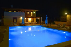Una piscina con sillas de descanso y sombrillas, un área de estar cubierta y un edificio con varias ventanas y un balcón.