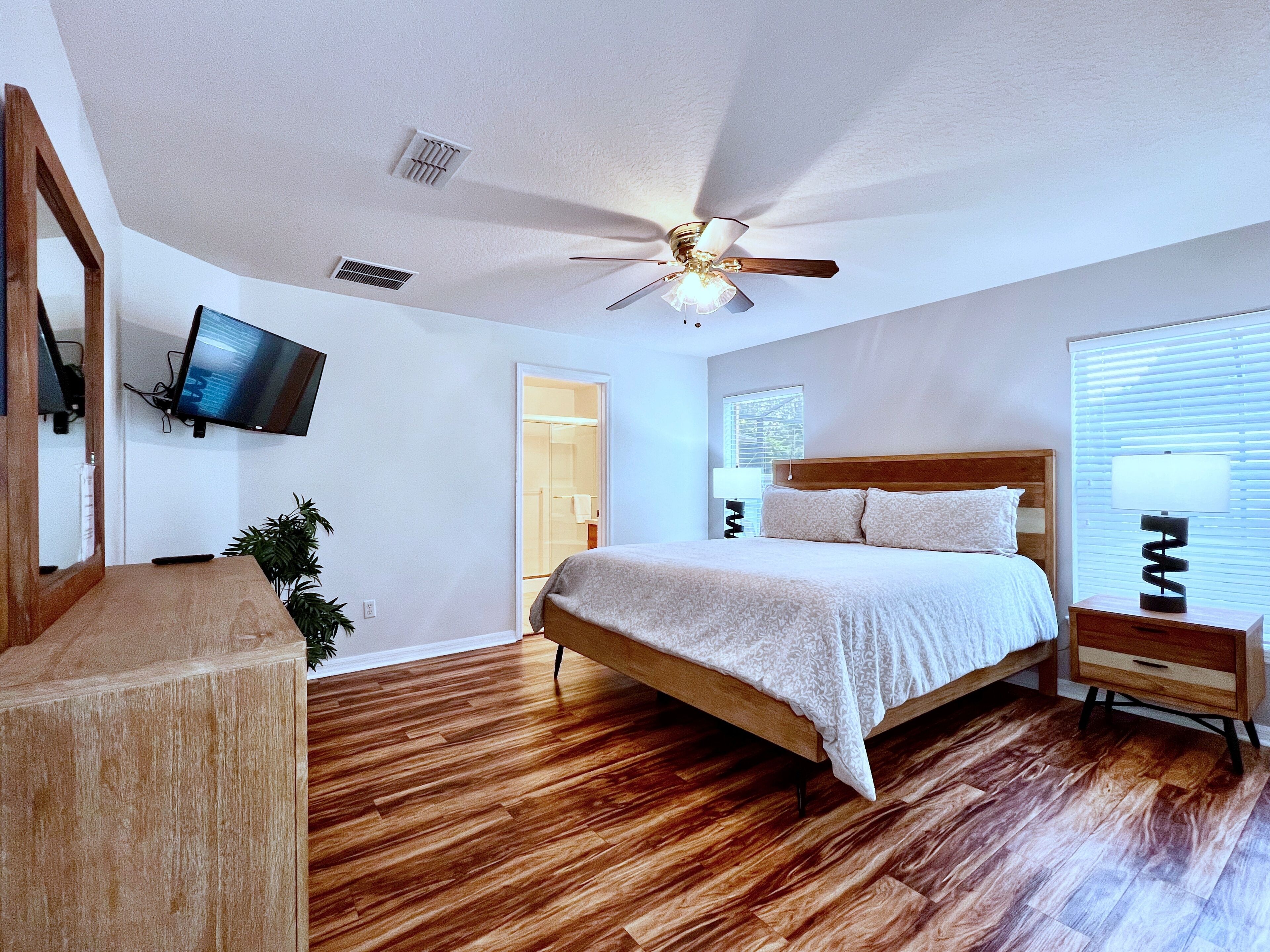 A bedroom with a wooden bed, a ceiling fan, a TV mounted on the wall, and a window with blinds.