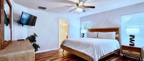A bedroom with a wooden bed, a ceiling fan, a TV mounted on the wall, and a window with blinds.