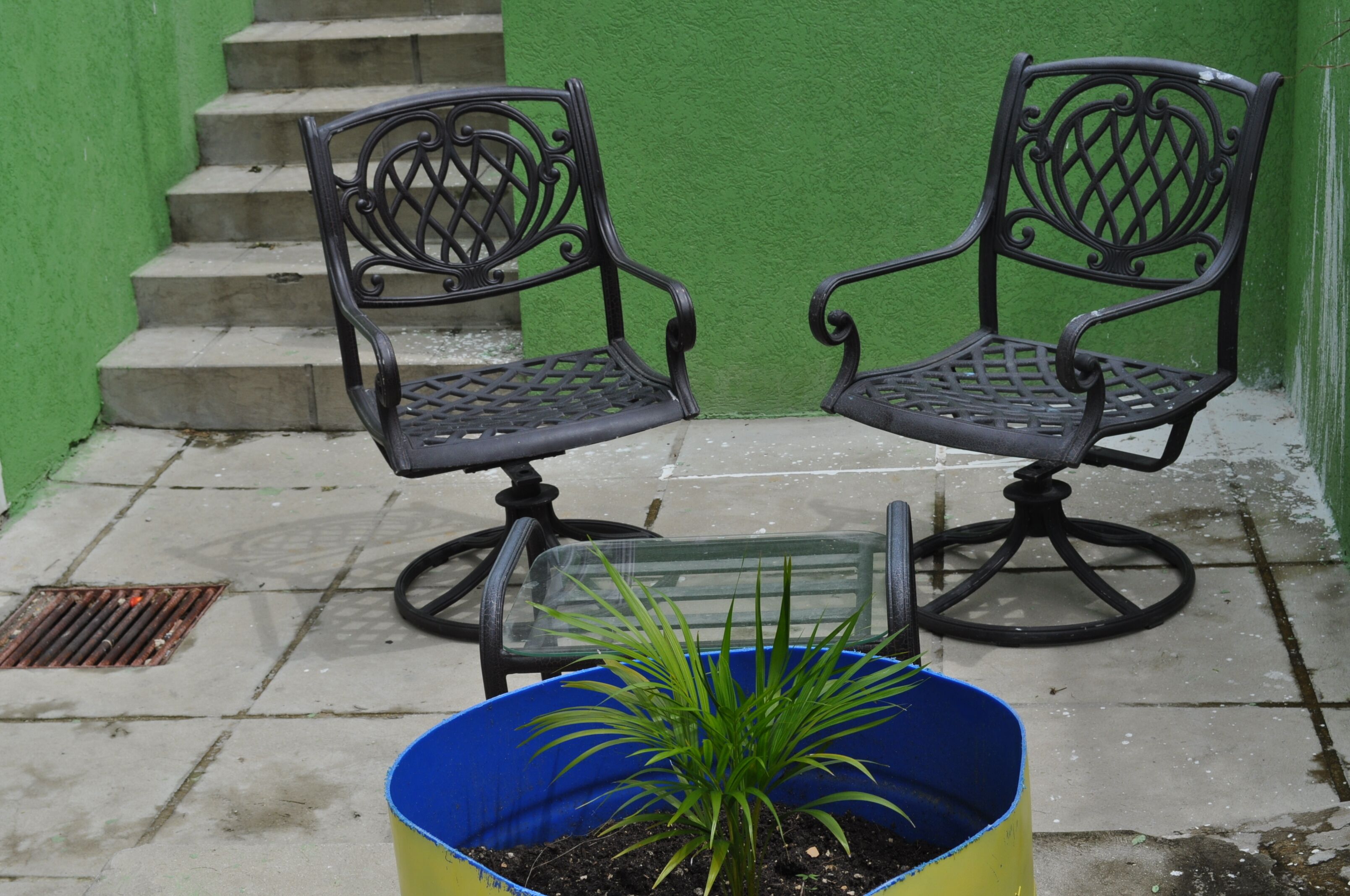 Two black metal chairs with intricate designs, placed on a tiled patio with a green wall and a set of stairs in the background.