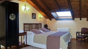 Quarto com teto inclinado, cama com cabeceira, mesa de cabeceira, uma cadeira, radiador e relógio na parede.