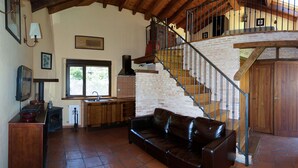 Interior rústico com escada de madeira, sofá de couro preto, área de cozinha e janela com vista para o verde.