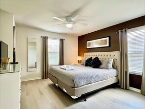 Une chambre à coucher comprenant un lit, un ventilateur de plafond, une table de chevet, une lampe et une fenêtre avec des stores.