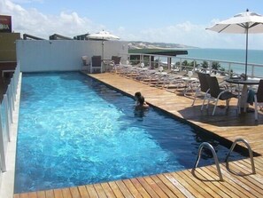 A swimming pool with a wooden deck, lounge chairs, and umbrellas. - Spacious apartment in Ponta Negra, 30m from the beach, balcony, oceanfront view. (Natal)