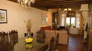 Un coin repas avec une table en bois, des chaises et un vase rempli de fleurs jaunes. On aperçoit un canapé et un escalier au fond.