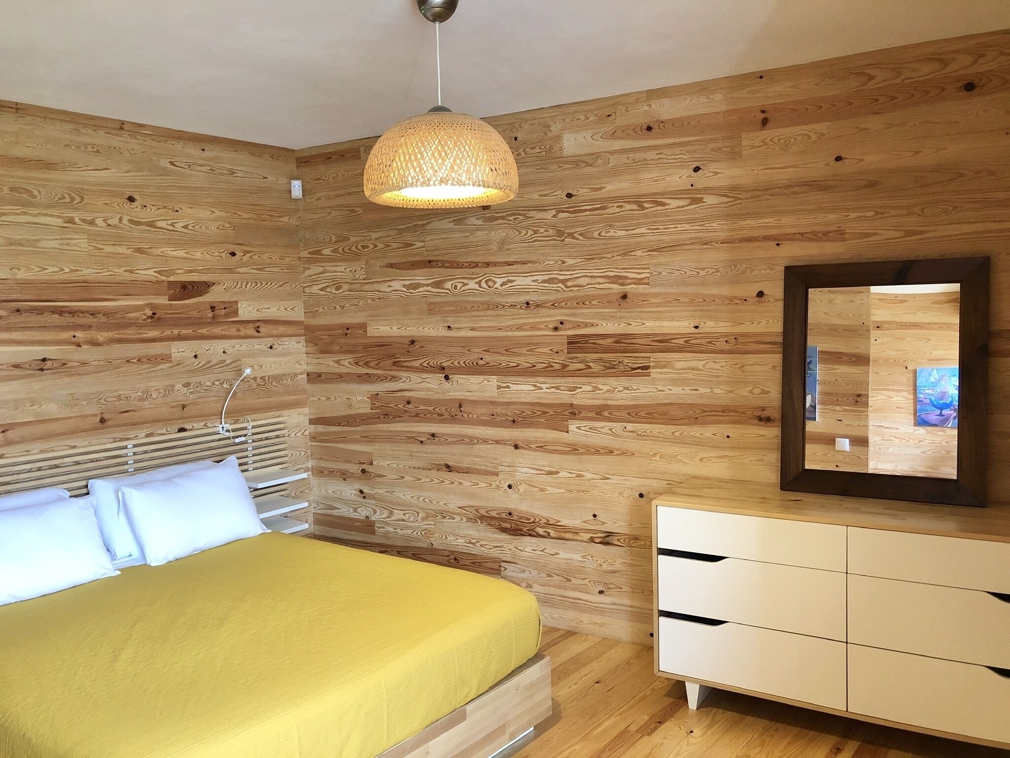 Une chambre avec un mur décoré de panneaux en bois, un lit avec des oreillers blancs, une couvre-lit jaune, une commode blanche et un luminaire suspendu.