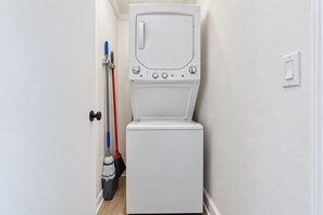 Een compact wit was- en droogcombitoestel aan de muur, in een smalle gang met een deur aan de ene kant en een lichtschakelaar op de muur.
