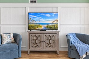Een televisie op een houten kast met een afbeelding van een strand, aan weerszijden geflankeerd door twee grijze fauteuils.