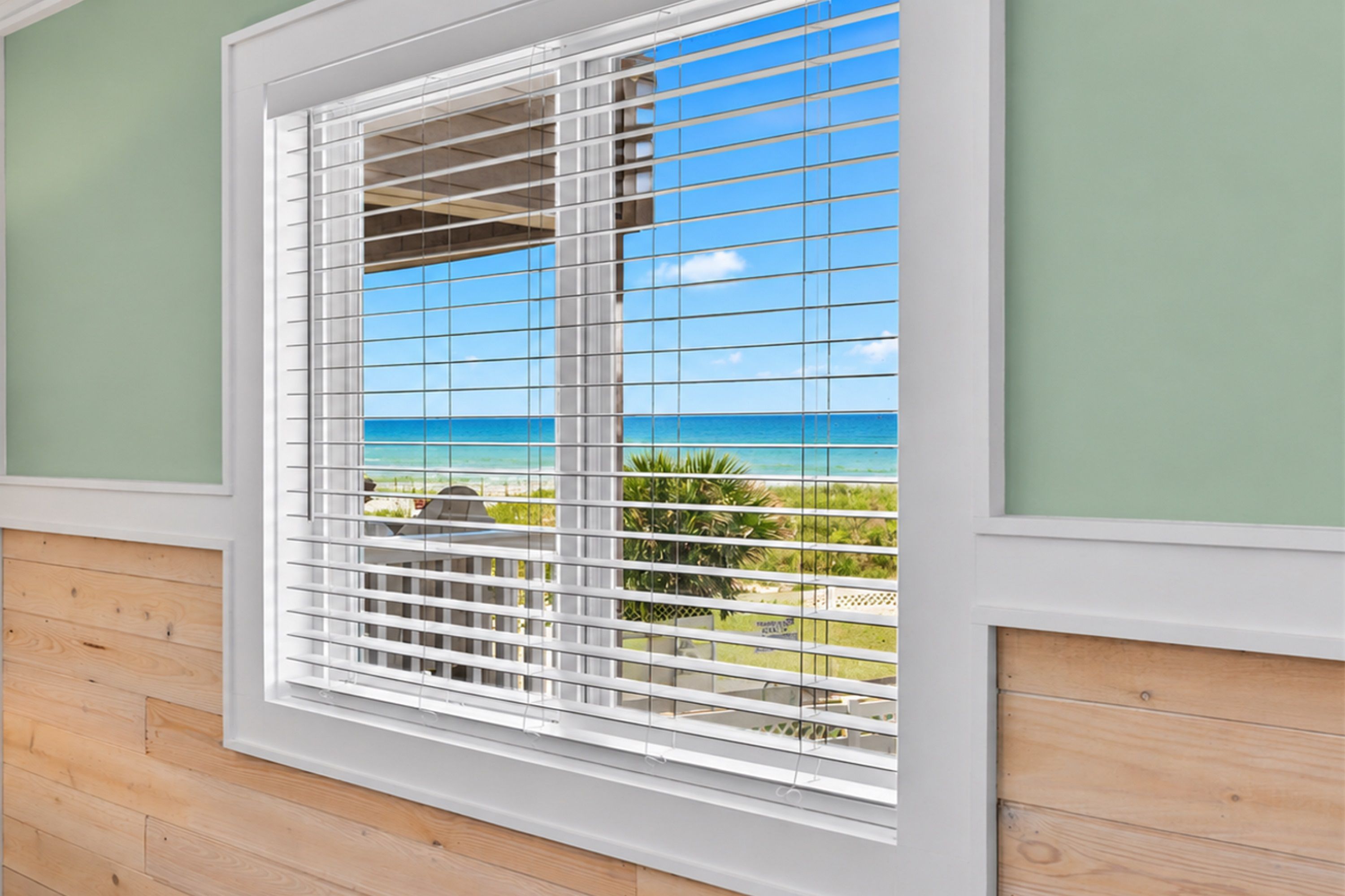 A window with horizontal blinds, a view of the ocean, and a wooden floor.