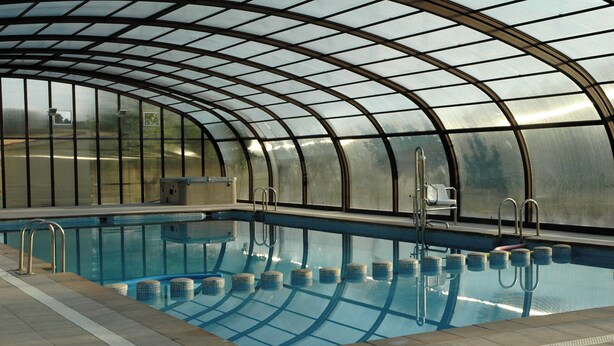An indoor swimming pool with a curved glass roof, clear water, and tiled steps leading into the pool.