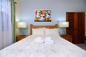 Cama bem arrumada com colcha estampada, duas luminárias de cabeceira, um painel de cabeceira de madeira e uma pintura abstrata colorida na parede.