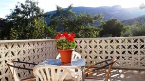 En terrasse med et bord og to stole, en potteplante med røde blomster, og udsigt til bjerge.