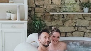 Un couple dans un bain à remous, avec un mur en pierre et des étagères ornées de plantes et de bougies.