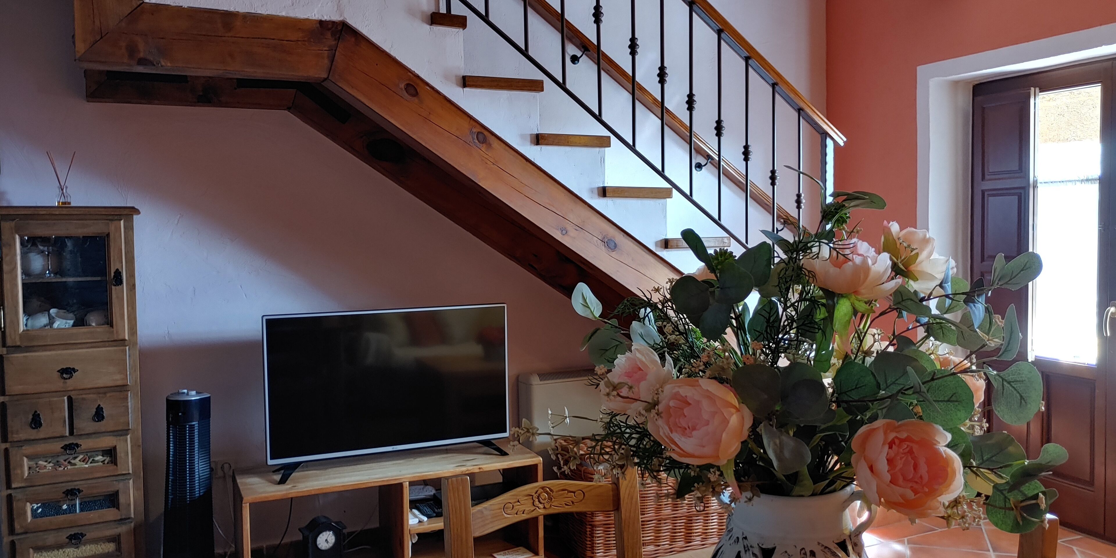 A living room with a TV on a wooden stand, a clock, a vase with flowers, and a staircase.