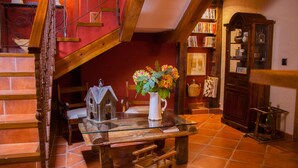Een rustiek interieur met een houten trap, een tafel met glazen blad, een vogelhuisje en een vaas, en een boekenplank vol boeken en decoratieve objecten.