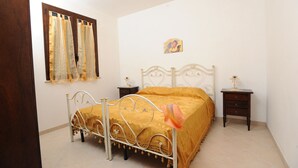 Un dormitorio con estructura de cama metálica, cubrecama amarillo, mesita de noche con lámpara y ventana con cortinas.