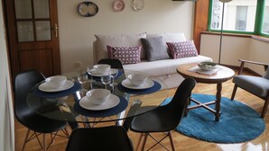 Ein Essbereich mit einem Glastisch, schwarzen Stühlen und Geschirr. Im Hintergrund befindet sich eine Couch mit gemusterten Kissen.
