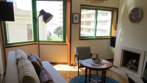 Ein Wohnzimmer mit einer Couch, einem Tisch mit einer Schale, einem Sessel, einem Kamin und einem Fenster mit Blick auf Gebäude.