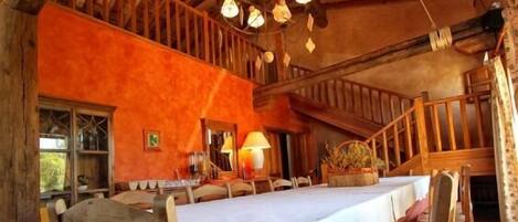A rustic dining area with a long table set for multiple people, wooden beams on the ceiling, and a warm, orange wall.