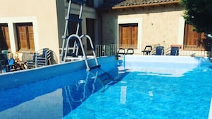 En swimmingpool med en stege, omgiven av stolar och en byggnad med trädörrar.