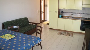 Una pequeña cocina con gabinetes amarillos, un lavamanos y una estufa. Hay una mesa de comedor con mantel azul y un sofá verde en la sala de estar.