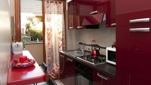 Eine kompakte Küche mit Gasherd, Mikrowelle und roten Schränken. Ein Fenster mit Vorhang ist sichtbar, und ein kleiner Tisch mit roter Kerze und Blumen steht davor.