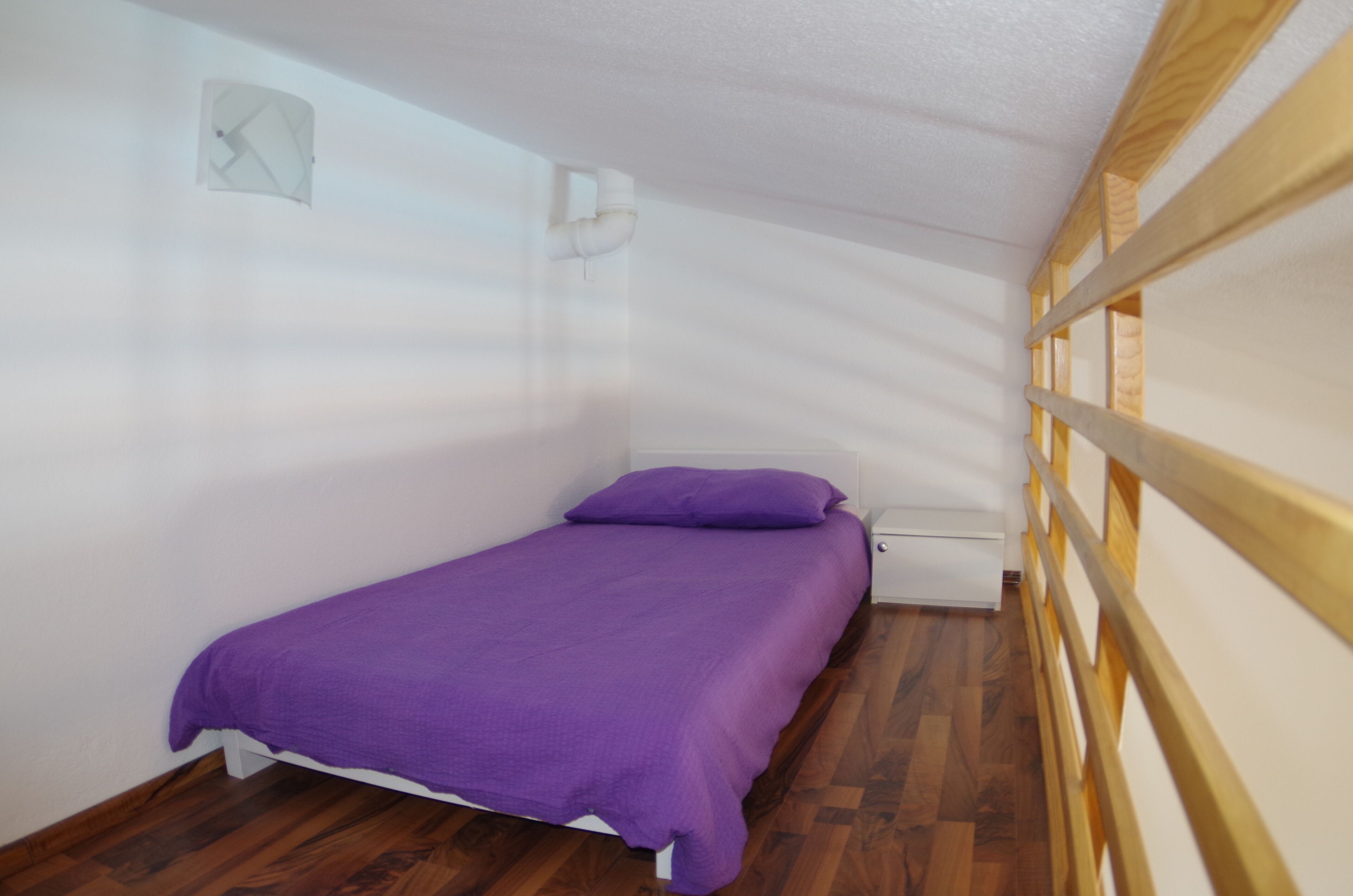 Pieni huone, jossa on yksi sänky, violetti sängynpeite, valkoiset seinät ja puulattia.