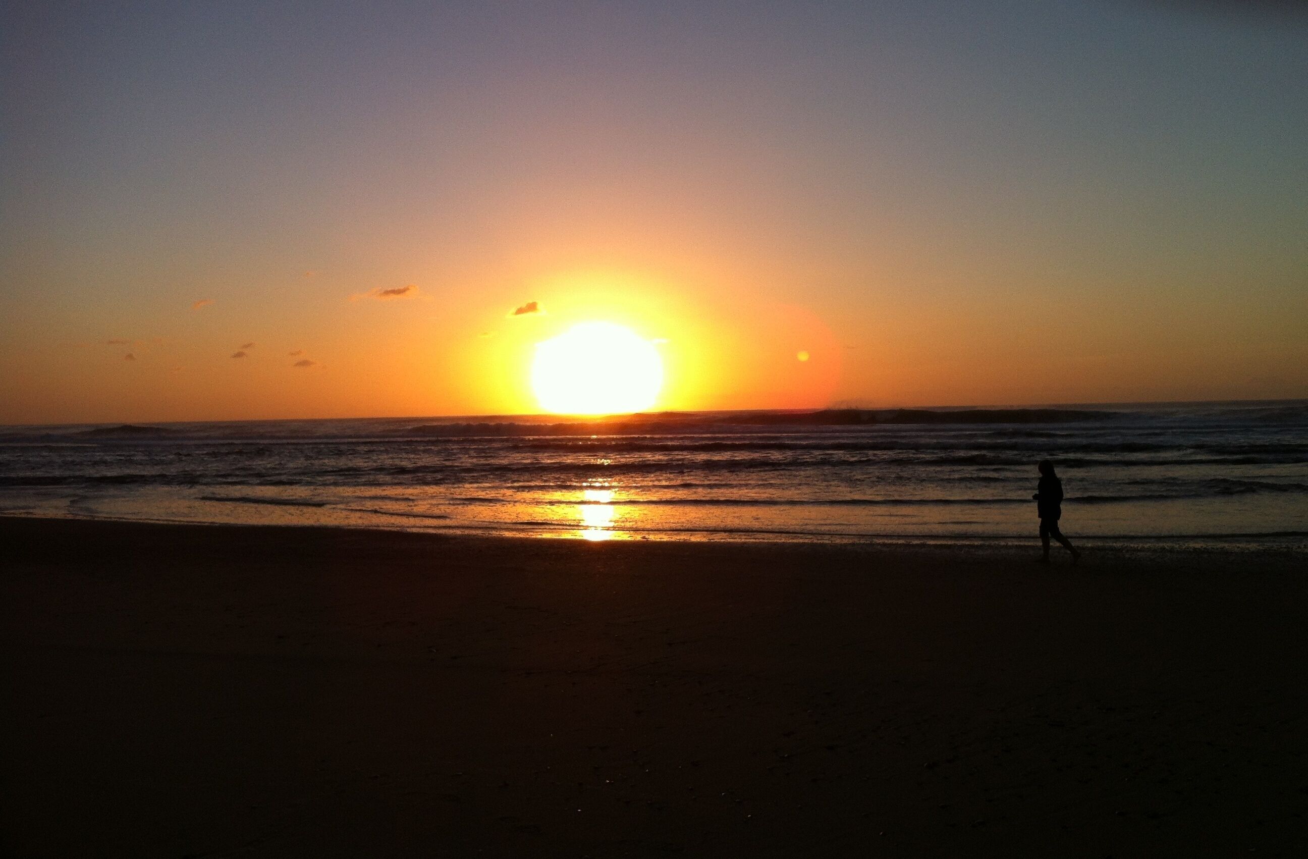 Un atardecer sobre una playa con una persona caminando.