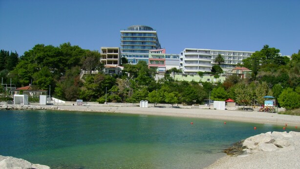 A coastal area with a pebble beach, clear water, and buildings in the background.
