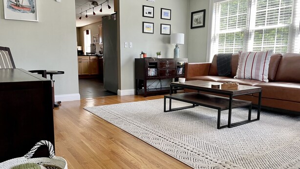 A living room with a sofa, coffee table, and a rug. There is a kitchen in the background with cabinets and a refrigerator. - Contemporary East Side Townhouse (Providence)